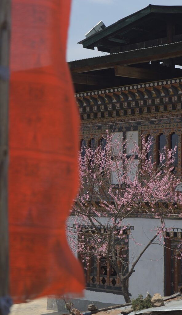Bandera de oración roja vertical junto a un arbol y una casa de estilo tradicional butanés (Bhutan)