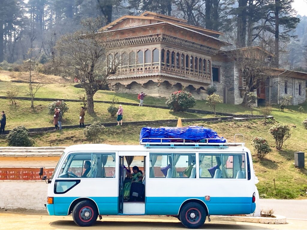 Bus/Taxi tradicional con visitantes del Dochula Pass en Bután (Bhuta). Al fondo una casa tradicional butanesa con detalles de madera.